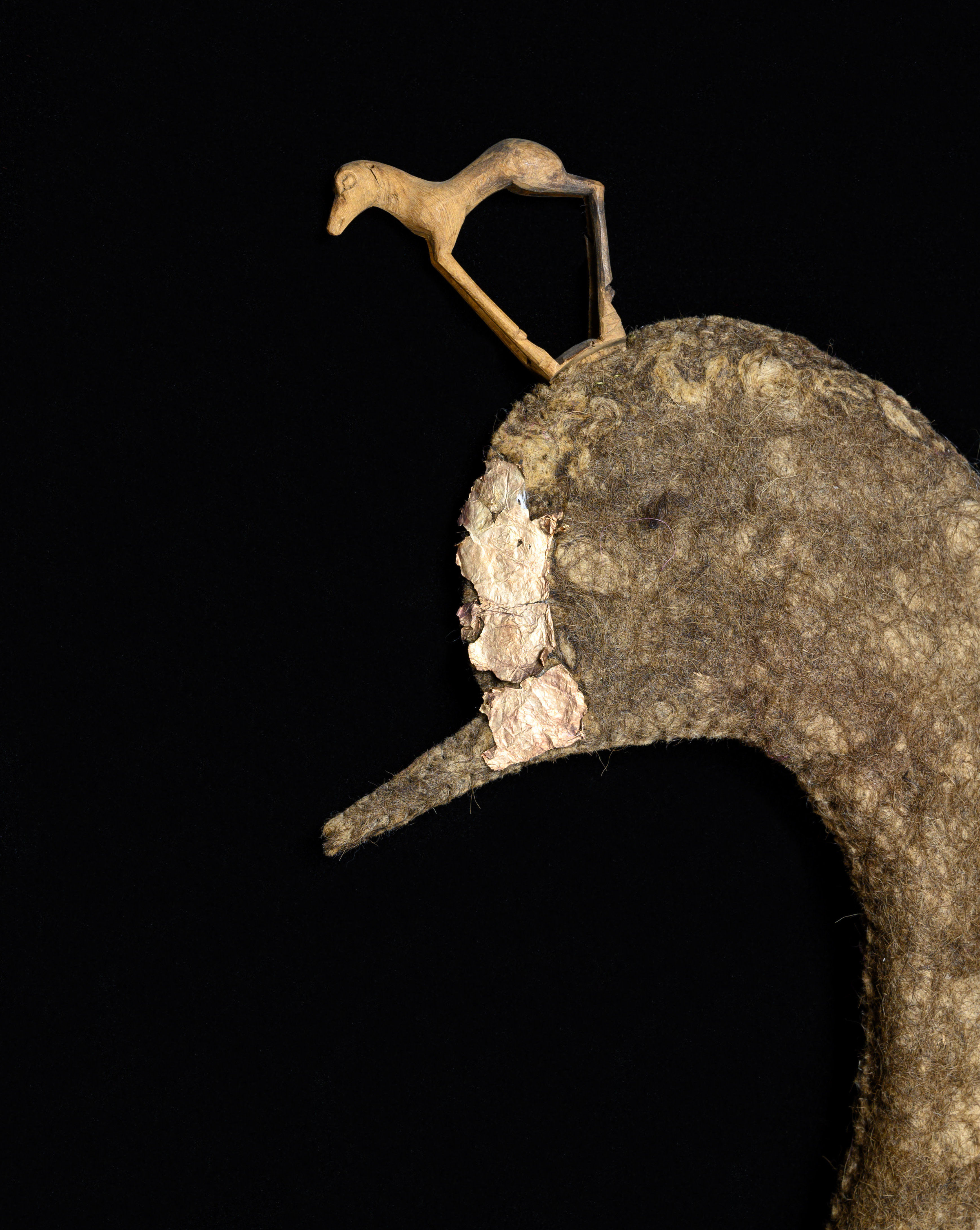 Das Bild zeigt einen Teil einer Filzmütze, die wie ein Entenkopf geformt ist, auf schwarzem Grund. Auf dem Entenkopf sieht man eine filigrane Holzfigur in Pferdedorm.