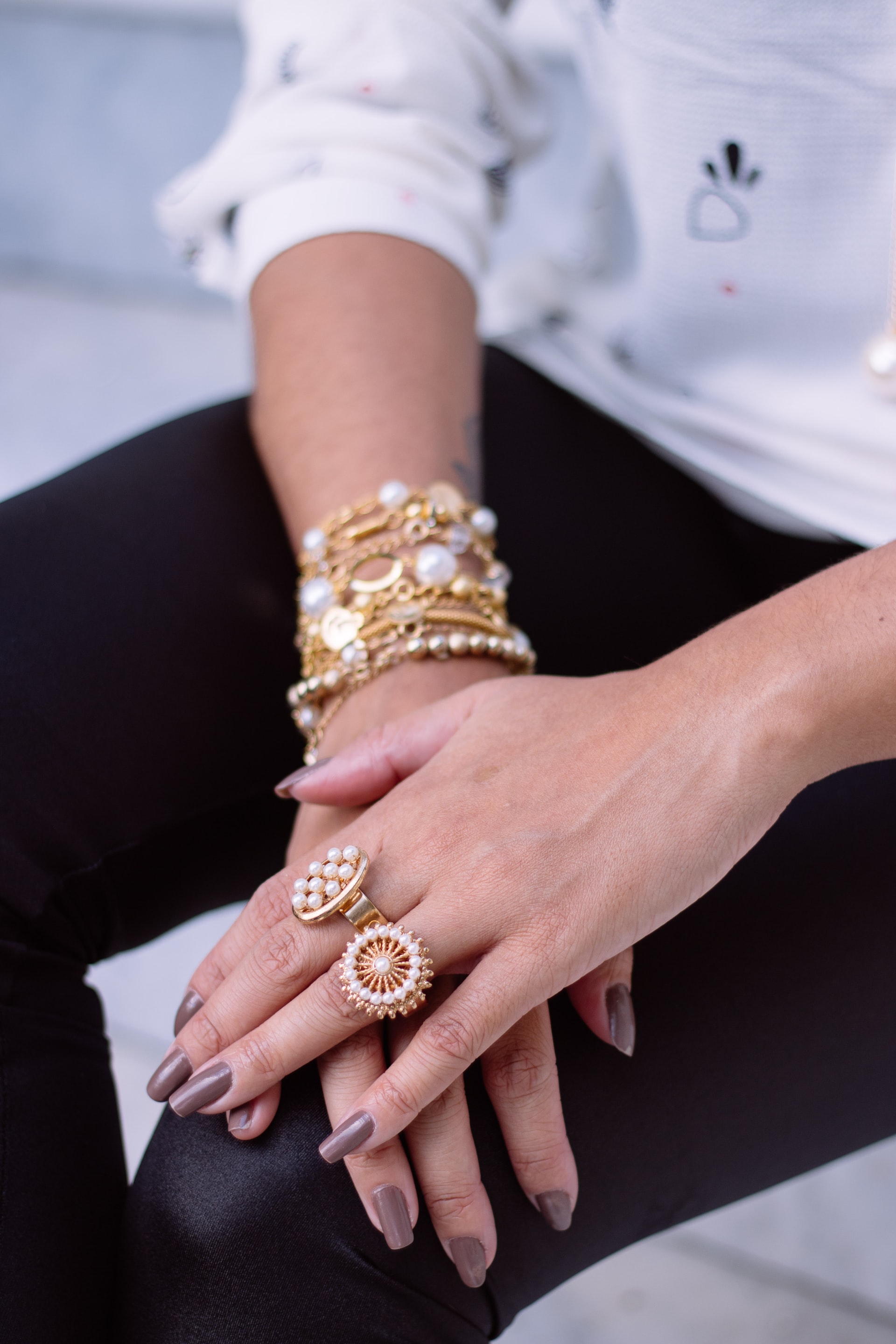 Zwei Hände auf einem Knie übereinandergelegt. Daran viele Ringe und Armbänder in Gold. Die Fingernägel sind hellbraun lackiert